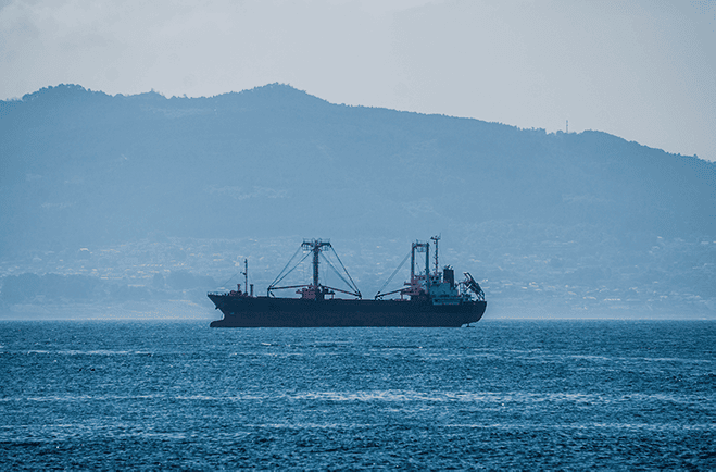 Large cargo ship at sea, its structure and cranes visible against a backdrop of a hazy mountain range, under the soft glow of either early morning or late afternoon light, symbolizing maritime transport and global trade.