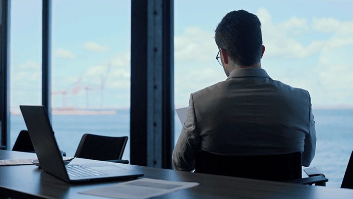 Business professional in a suit working on a laptop at a conference table with an ocean view through floor-to-ceiling windows, symbolizing focused work environment and corporate success.