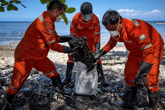 Three individuals in orange hazmat suits collecting black oily substances from a beach, with a clear blue sky and calm sea in the background, symbolizing an environmental cleanup effort on a coastal area.
