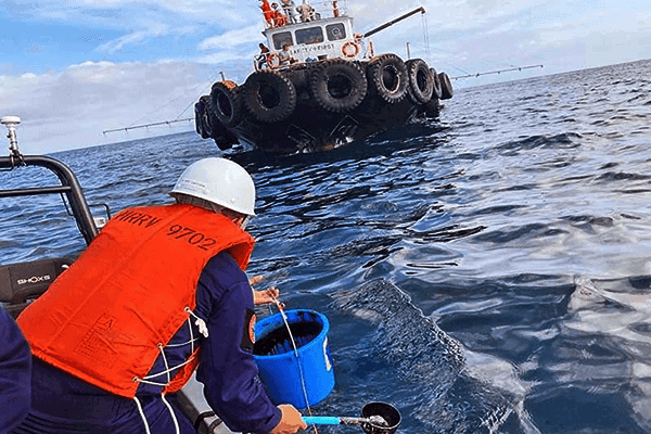 Offshore technician in safety helmet collecting water samples from the ocean aboard a small boat, with a large multi-tiered amphibious vehicle in the background, under a partly cloudy sky, symbolizing marine research and exploration.