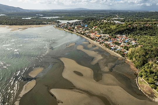 Aerial view of a coastal village with distinct sand patterns in the shoreline, surrounded by lush greenery and a cloudy sky, showcasing the unique interaction between human settlement and natural landscape.