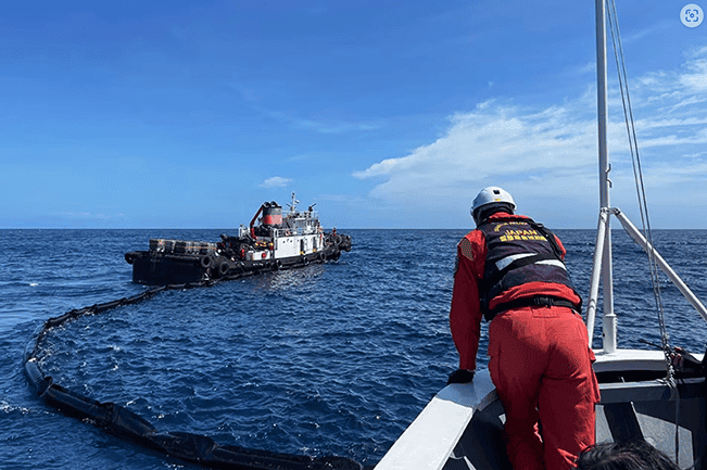 A marine cleanup crew at work in the open ocean, with a person in a red and yellow uniform observing a vessel towing a long floating barrier designed to collect trash, under a clear blue sky and calm sea, symbolizing environmental effort.