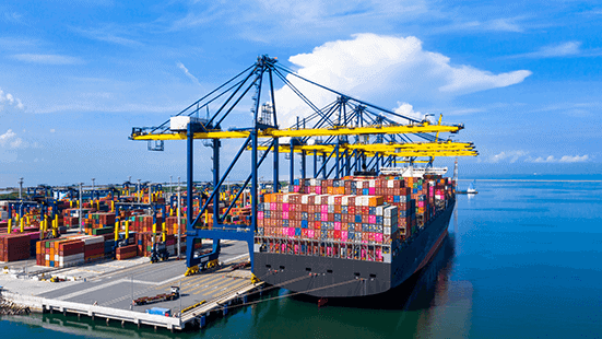 A large cargo ship loaded with colorful containers docked at a commercial port, under a clear blue sky and calm ocean waters, symbolizing global maritime trade and logistics.