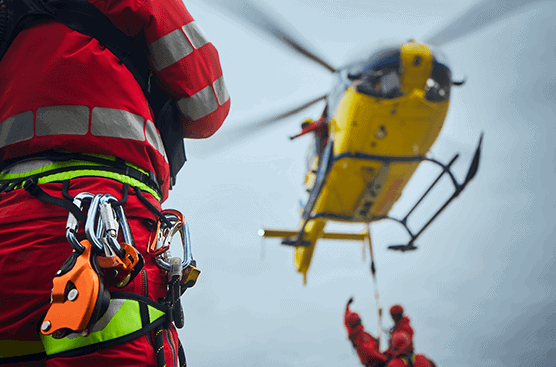 Close-up view of a rescue worker in a bright red suit with safety harnesses, with a yellow rescue helicopter and another suspended rescue worker in the background, symbolizing aerial rescue operations.