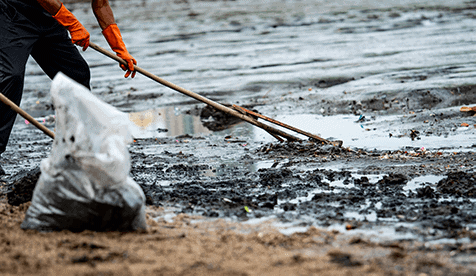 Individual wearing orange gloves, using a long stick to drag a black garbage bag across a muddy terrain littered with patches of oil, symbolizing environmental cleanup efforts